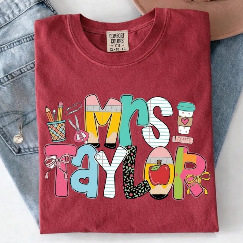 May include: A brick red t-shirt with a graphic print featuring the text Mrs. Taylor. The design includes school-themed illustrations such as a pencil cup, scissors, a paperclip, a coffee cup, a red apple, and bows in shades of yellow, blue, and pink.