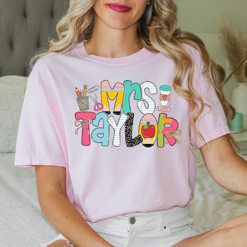 May include: A light pink short-sleeve t-shirt featuring a colorful graphic design with the text Mrs. Taylor. The design includes illustrations of pencils in a cup, scissors, a coffee cup with a heart, and a red apple, all in various patterns.