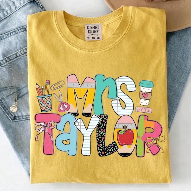 May include: Yellow Comfort Colors t-shirt with the name "Mrs. Taylor" in colorful, playful lettering. The design includes school-themed graphics like pencils, scissors, and a coffee cup. The shirt is folded on a pair of blue jeans.