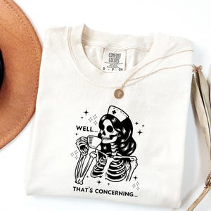 May include: Cream-colored t-shirt featuring a black and white illustration of a skeleton in a nurse's cap, drinking from a mug. The text "WELL... THAT'S CONCERNING..." is below the graphic. A gold necklace and a brown hat are also visible.