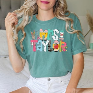 May include: A teal short-sleeve t-shirt featuring a colorful graphic print with the text "Mrs. Taylor". The design includes school-themed illustrations such as a pencil cup, scissors, a red apple, and a coffee cup, all rendered in various patterns and bright colors.