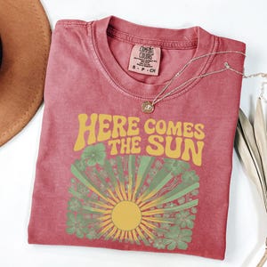 May include: A dusty rose short-sleeve t-shirt featuring the text "HERE COMES THE SUN" in yellow, arched above a graphic of a yellow sun with green rays and green flowers. The shirt has a Comfort Colors brand label at the inner neckline.