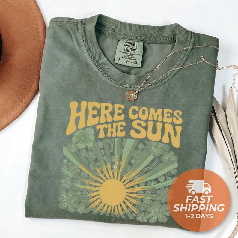 May include: An olive green short-sleeve t-shirt with a crew neckline. The shirt features a graphic print with the yellow text "HERE COMES THE SUN" above a stylized sunburst design surrounded by green floral illustrations on the front chest.
