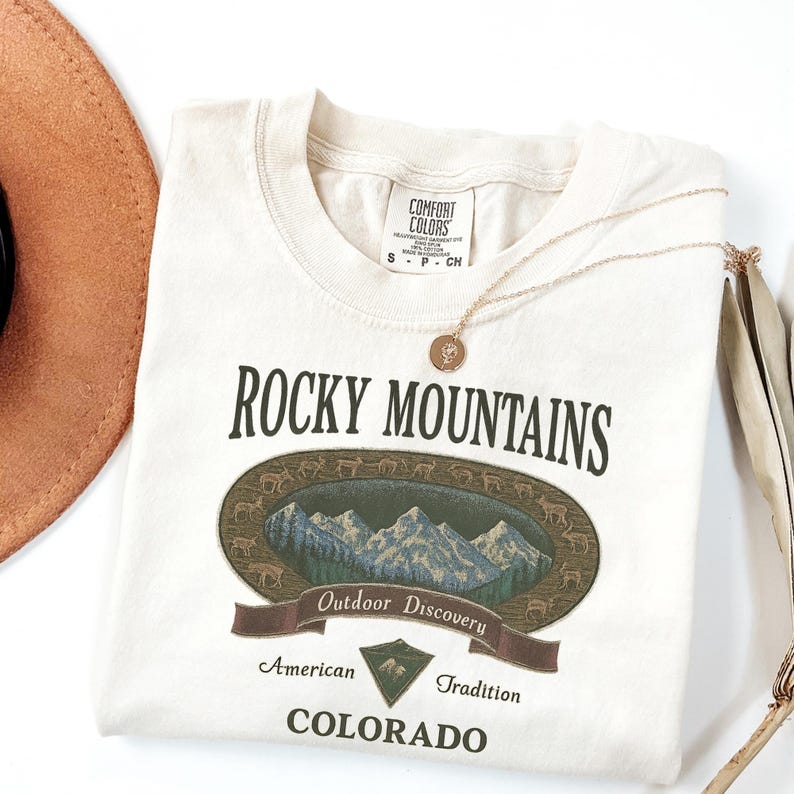 Pu&ograve; includere: T-shirt color crema con grafica "Rocky Mountains". Il design include un'immagine ovale di montagne, alberi e cervi, con il testo "Outdoor Discovery", "American Tradition" e "Colorado". Sono visibili anche una collana d'oro e un cappello marrone.
