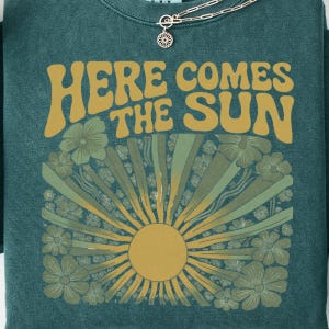 May include: A dark teal crewneck sweatshirt featuring the text "HERE COMES THE SUN" in a bold, mustard yellow font. Below the text is a graphic of a sun with radiating beams surrounded by stylized flowers in shades of green and yellow.