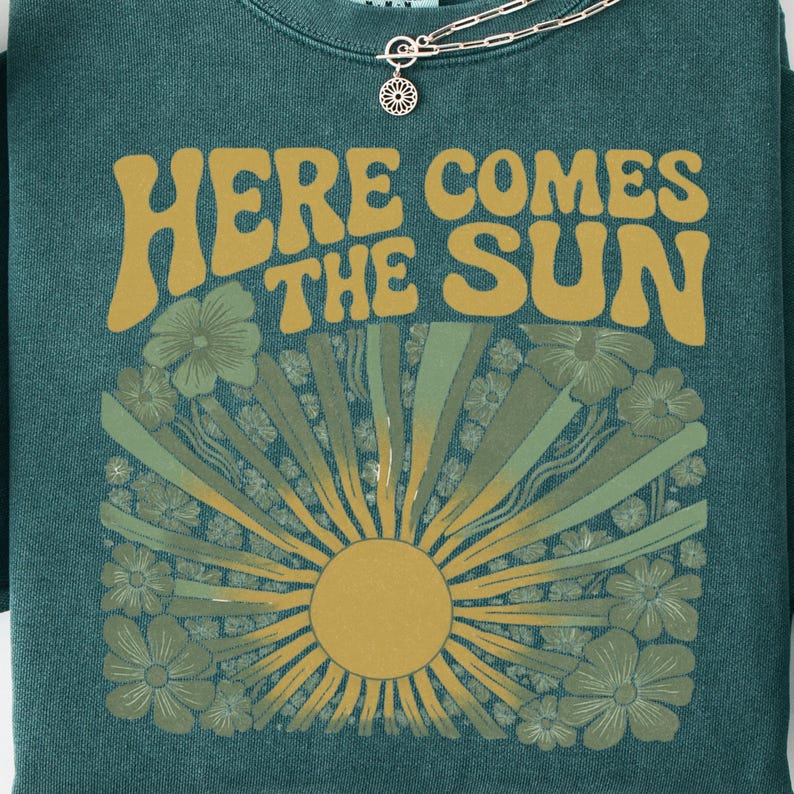 May include: A dark teal crewneck sweatshirt featuring the text "HERE COMES THE SUN" in a bold, mustard yellow font. Below the text is a graphic of a sun with radiating beams surrounded by stylized flowers in shades of green and yellow.