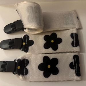 Puede incluir: Tres pinzas para chupete de tela blanca con estampados de flores negras y pinzas de plástico negro. Una pinza tiene cierre de velcro. El diseño floral tiene un centro amarillo. Las pinzas están diseñadas para sujetarse a la ropa del bebé.