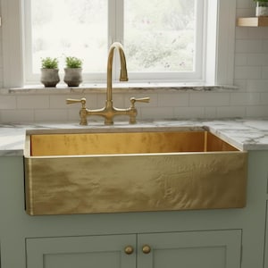May include: A gold-coloured farmhouse sink is the focal point of this kitchen. The sink is set into a light green cabinet with gold hardware. A gold tap is mounted above the sink. The countertop is white with grey veining. A window is in the background.