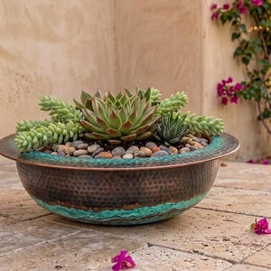May include: A round, hammered copper bowl planter with a wide rim and a teal patina finish at the base. The bowl is filled with soil, small smooth river stones, and a variety of green succulents, including a central rosette-shaped plant.