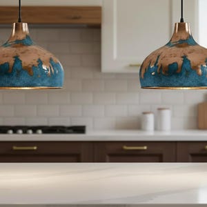 May include: Two pendant lights with copper-colored tops and teal blue bottoms hang in a kitchen. The lights are suspended above a white countertop and dark brown cabinets. The kitchen has white tile and a wooden range hood.