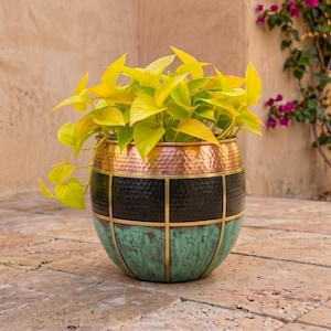 Könnte beinhalten: Ein runder Metall-Pflanztopf mit gehämmertem Muster, bestehend aus drei horizontalen Bändern in Kupfer, Schwarz und einer verwitterten türkisfarbenen Patina. Der Topf ist durch dünne vertikale und horizontale messingfarbene Linien unterteilt.