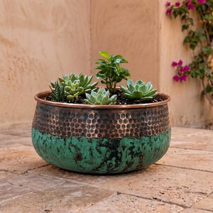 May include: A round, bowl-shaped planter featuring a hammered copper upper section and a textured, weathered teal green lower base. The planter is filled with dark soil and several small, green succulent plants of varying shapes and sizes.