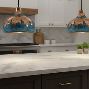 May include: Two pendant lights with copper and teal finishes hang above a kitchen island. The lights have a unique, textured design. The kitchen features white cabinets, a marble countertop, and a dark brown island base.
