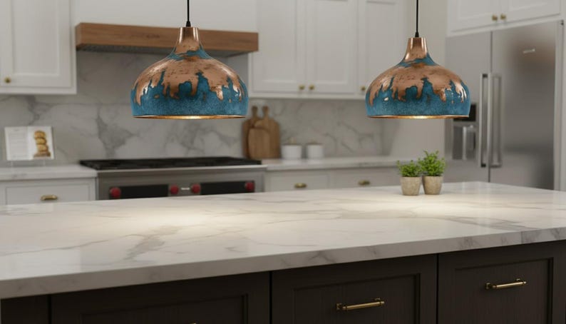 May include: Two pendant lights with copper and teal finishes hang above a kitchen island. The lights have a unique, textured design. The kitchen features white cabinets, a marble countertop, and a dark brown island base.
