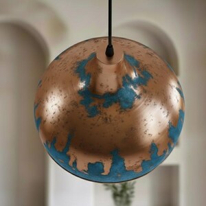 May include: A copper-colored pendant light fixture with a globe shape. The surface has a distressed finish with patches of turquoise-blue. The light hangs from a black cord, suitable for interior lighting.