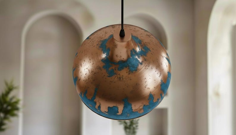 May include: A copper-colored pendant light fixture with a globe shape. The surface has a distressed finish with patches of turquoise-blue. The light hangs from a black cord, suitable for interior lighting.