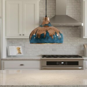 May include: A pendant light fixture with a copper-colored top and a blue and brown glazed bottom hangs in a kitchen. The kitchen features white cabinets, a stainless steel refrigerator, and a stove. The countertop is a light-colored granite.