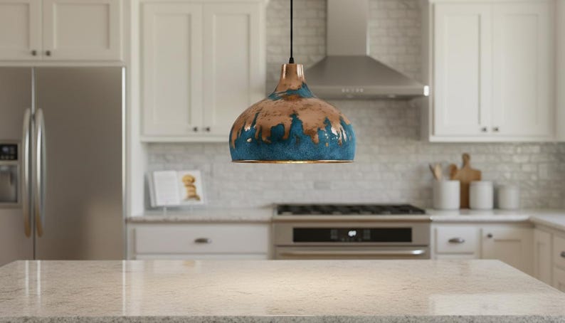May include: A pendant light fixture with a copper-colored top and a blue and brown glazed bottom hangs in a kitchen. The kitchen features white cabinets, a stainless steel refrigerator, and a stove. The countertop is a light-colored granite.