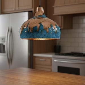 May include: A pendant light fixture with a brown and blue glazed finish hangs in a kitchen. The light has a dome shape with a gold interior. The kitchen features wooden cabinets, a stainless steel refrigerator, and a wooden countertop.