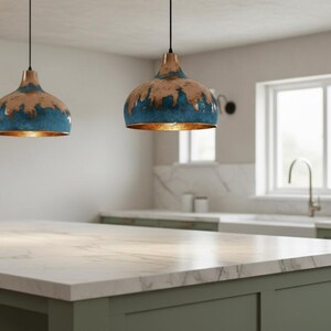 May include: Three pendant lights with a blue and brown finish hang above a kitchen island. The lights have a metallic gold interior. The kitchen features a marble countertop and green cabinets. A window and plants are visible in the background.