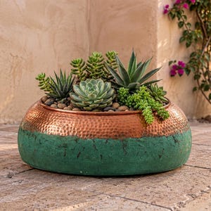 May include: A round, low-profile planter featuring a hammered copper upper section and a textured, matte green lower base. The planter is filled with a variety of green succulents and small, smooth river stones arranged in the soil.