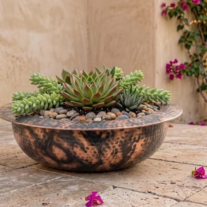 Pode incluir: Uma taça redonda de cobre martelado com uma borda larga, preenchida com uma variedade de suculentas verdes e pequenas pedras de rio lisas e multicoloridas. A superfície metálica apresenta uma pátina escura e manchada com tons de cobre.