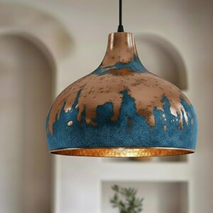 May include: A copper-colored pendant light with a blue and brown distressed finish. The light fixture has a bell-shaped shade and a gold-colored interior. The light hangs from a black cord, suitable for home decor.