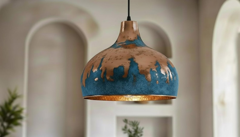May include: A copper-colored pendant light with a blue and brown distressed finish. The light fixture has a bell-shaped shade and a gold-colored interior. The light hangs from a black cord, suitable for home decor.