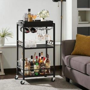 May include: A black metal bar cart with three shelves and wheels. The top shelf holds a tray with a floral arrangement and cocktail glasses. The middle shelf has wine glasses and a decorative container. The bottom shelf displays various liquor bottles.