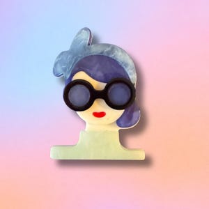 May include: A whimsical brooch featuring a stylized face with dark-rimmed glasses, purple hair, and red lips. The brooch has a light green base and is set against a gradient background of pink and blue.
