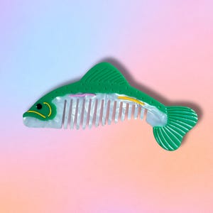 May include: A green fish-shaped hair comb with a white and pearlescent body. The comb has a yellow eye, pink and yellow accents, and a green tail fin. The comb's teeth are white. The background is a gradient of pink, blue, and purple.