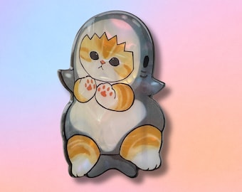 May include: A whimsical cat-shaped pin, featuring a kitten in a shark costume. The pin has a glossy finish with a gradient background. The kitten is orange and white, with a gray shark costume. The pin is approximately 2 inches tall.