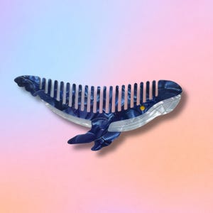 May include: A whale-shaped hair comb in shades of blue and white. The comb features a marbled pattern on the whale's body and a row of white teeth. A small yellow accent is visible on the whale's eye.