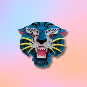 May include: A blue tiger head brooch with black stripes, yellow whiskers, and a pink mouth. The brooch has a glittery texture and is set against a gradient background of pink and blue.