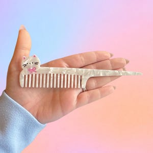 May include: A white cat-shaped comb with a pink bow and black details. The comb has a pearlescent finish and a long, pointed handle. The comb is held in a hand against a pastel gradient background.
