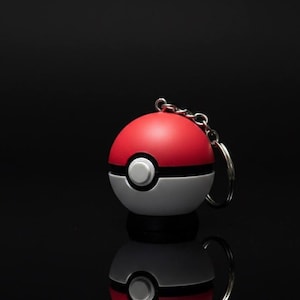 Può includere: Un portachiavi Poké Ball rosso e bianco con una catena e un anello argentati. La palla ha una striscia nera e un bottone bianco al centro. Il portachiavi è su una superficie nera riflettente.