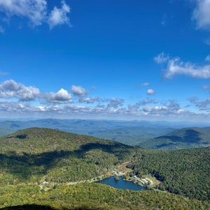 May include: A panoramic view from a mountaintop, showcasing a lush green valley with a small lake in the center. The sky is a vibrant blue with fluffy white clouds.