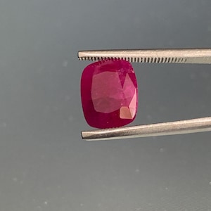Peut inclure: Une pierre précieuse rubis taille coussin avec une couleur rouge profond et saturé et de multiples facettes qui reflètent la lumière. La pierre est maintenue par une pince en métal argenté, mettant en valeur sa qualité translucide.