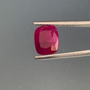 Op de afbeelding: Een robijn edelsteen in kussenslijpvorm met een diepe, verzadigde rode kleur en meerdere facetten die het licht reflecteren. De edelsteen wordt stevig vastgehouden door een zilverkleurige metalen pincet, wat de doorschijnende kwaliteit toont.