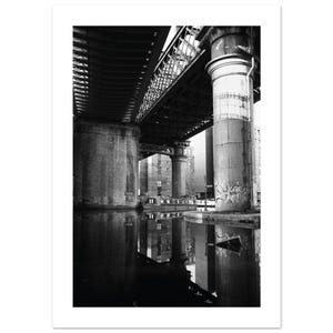 May include: Black and white photograph of a bridge over water. The underside of the bridge is visible, with support columns and metal beams. The water reflects the bridge and surrounding buildings. A boat is visible in the water.