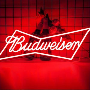 Puede incluir: Un letrero de neón rojo con el logotipo de Budweiser en blanco. Se ven dos botellas de cerveza detrás del letrero, con flores entre ellas. El fondo es un patrón de cuadrícula roja.