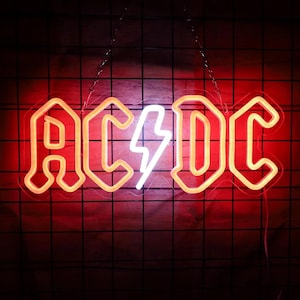 May include: A neon sign displaying the letters "AC/DC" in a bold, outlined font, with a white lightning bolt replacing the slash. The sign glows with a warm orange and red light against a dark background.