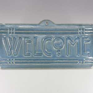 May include: A rectangular, light blue ceramic sign with the word "WELCOME" in raised, white lettering. The sign has a small hole at the top for hanging and decorative lines. The sign is a welcoming home decor item.