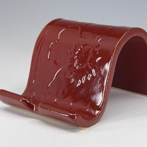 May include: A burgundy ceramic book stand with a curved design. The stand features an embossed floral pattern on the front. The stand is designed to hold a book or tablet. The stand is made of ceramic.
