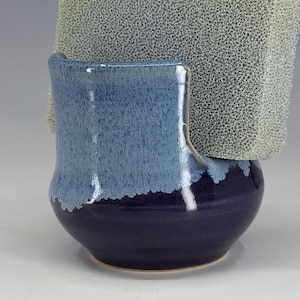 May include: A ceramic sponge holder with a two-tone blue glaze. The top portion of the holder is a lighter blue, transitioning to a darker blue at the base. A textured, light green sponge is placed in the holder.