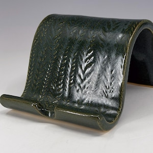 May include: A dark green ceramic phone stand with a glossy finish and a textured leaf pattern embossed across the curved surface. The stand features a wide base with a small front lip designed to hold a mobile device in a horizontal position.
