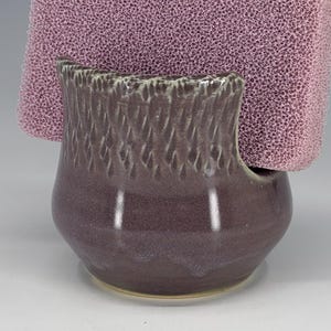 May include: A handcrafted, dark purple ceramic sponge holder with a textured exterior and a light tan rim. A pink sponge is placed inside the holder. The holder has a curved design and a smooth, glossy finish.