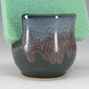 May include: A handmade ceramic sponge holder with a dark green base, transitioning to blue and burgundy hues. The holder has a curved shape and holds a green sponge. The glaze has a speckled appearance.