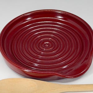 May include: A deep red ceramic spoon rest with a circular, ridged design. The spoon rest has a curved edge and a small indentation for holding a spoon. The wooden spoon is partially visible in the image.