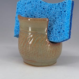 May include: A handmade ceramic sponge holder in a muted green and brown glaze. The holder has a textured, carved design and holds a bright blue sponge. The holder is designed to sit on a countertop.
